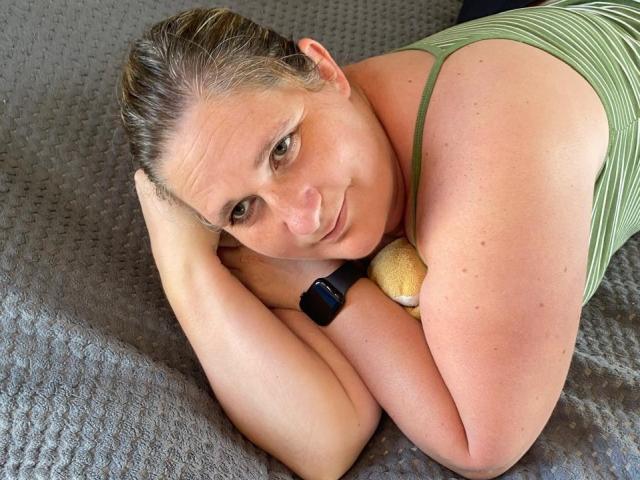 LiebeMaus is a curvy woman with small tits, lying on a bed, wearing a green striped tank top, black smartwatch, and holding a yellow ball, smiling at the camera.