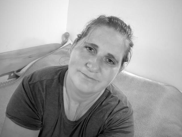 Black and white photo of LiebeMaus with wet, messy hair, wearing a dark shirt, smiling slightly, sitting on a textured surface with a skateboard in the background.