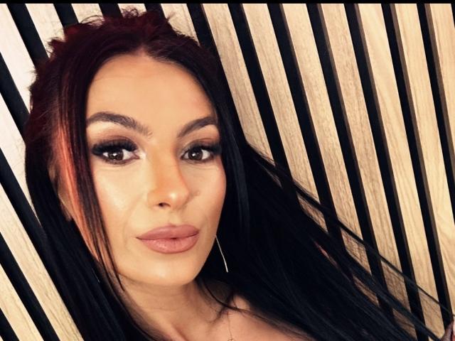 KinkyAmelie has red-black hair, full eyebrows, smoky eyes, thick eyelashes, glossy pink lips, and a subtle tan. She's wearing thin hoop earrings, looking directly at the camera.