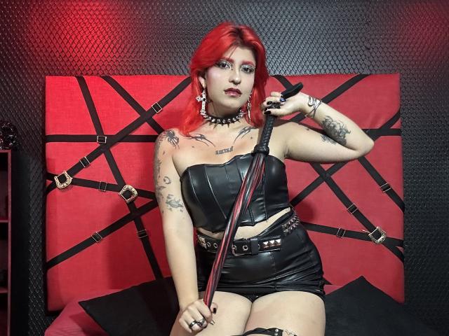 AthenaGrant with red hair, tattoos, black leather outfit, holding a red-and-black whip, sitting on a red and black bed with straps, wearing a spiked choker.