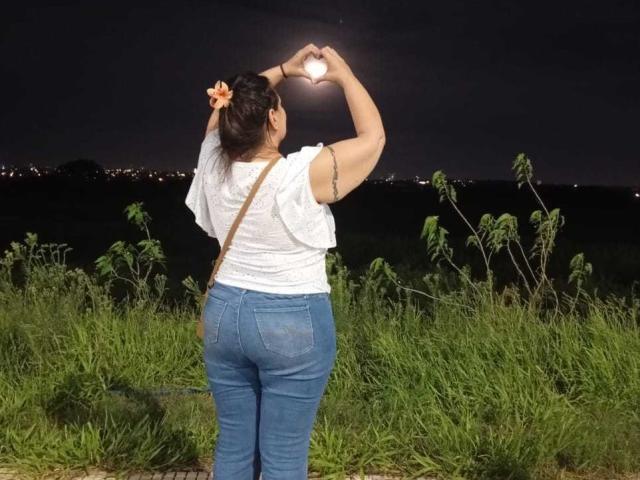 LadySelene with her back to the camera, making a heart shape with her hands, wearing a white shirt, blue jeans, and an orange flower in her hair, standing in grass.