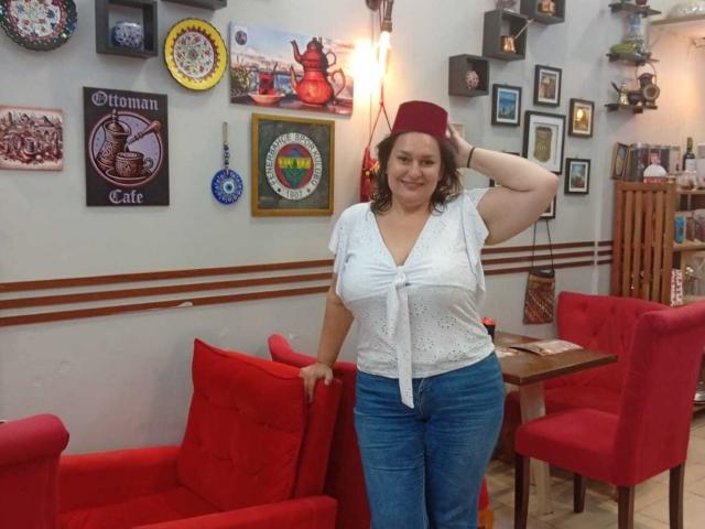 LadySelene with big boobs, wearing a white tied-up top, blue jeans, and a red hat, stands in a room with red chairs, smiling, hand on head.