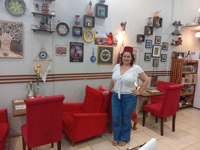 LadySelene with medium tits, white tied-up top, blue jeans, red headband, standing in room with red chairs, wall decorations, and wooden shelves, smiling, hands on hips.