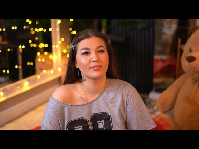 AkaliLuna with pouty lips, wearing a gray off-shoulder tee, brown hair in clips, subtle makeup, small tits, necklace, soft light, toy bear on right, warm glow.