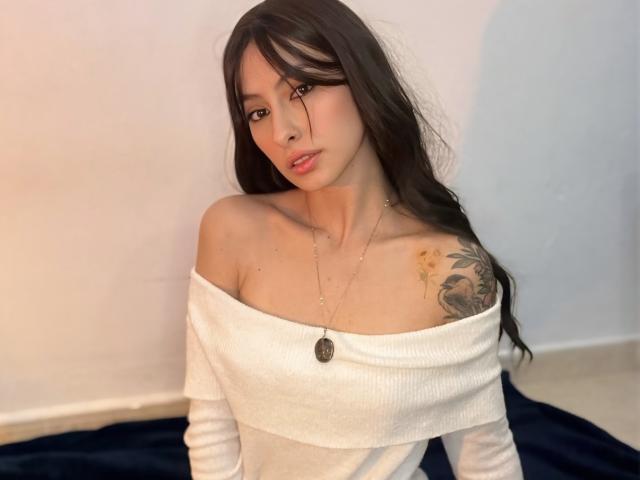 CamilleLoan with long dark hair, off-shoulder white sweater, small tits, tattoo on right shoulder, necklace, soft expression, sitting on dark blue rug, white wall in background.