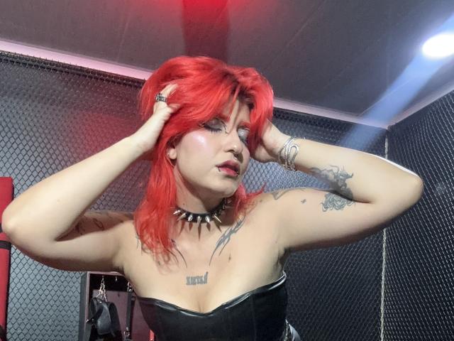 AthenaGrant met rood haar, handen in haar haar, gekleed in een zwarte bustier zonder bandjes, een choker, met tatoeages en een neusring, in een kamer met rode en blauwe lichten..