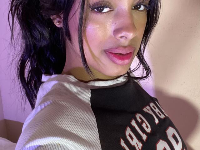 Melli69 with shiny skin, dark wavy hair, and pink lipstick, wearing a black and white sports shirt, looking at the camera with a slight smile, showing her pouty lips.