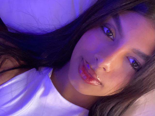 Melli69 has shiny purple lips and long black hair, lying on a white pillow with blue lighting. Her brown eyes reflect the light, and she's wearing a white shirt.