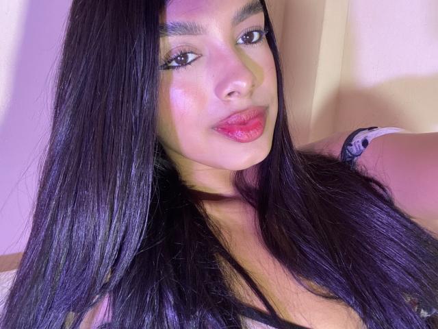 Melli69 has long, straight black hair, glossy pink lips, and a shiny face with subtle makeup. She's wearing a black lace top, showing her boobs. Purple light highlights her.