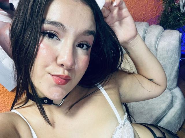 GagingZhara with long brown hair, pink lips, white lace bra, black choker, and a cute smile, posing with hand up, showing small tits and a slight tan.