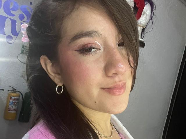 GagingZhara has pink eyeliner, shiny skin, and a subtle smirk. She's wearing pink, hoop earrings, and has brown hair with purple highlights, looking cute.