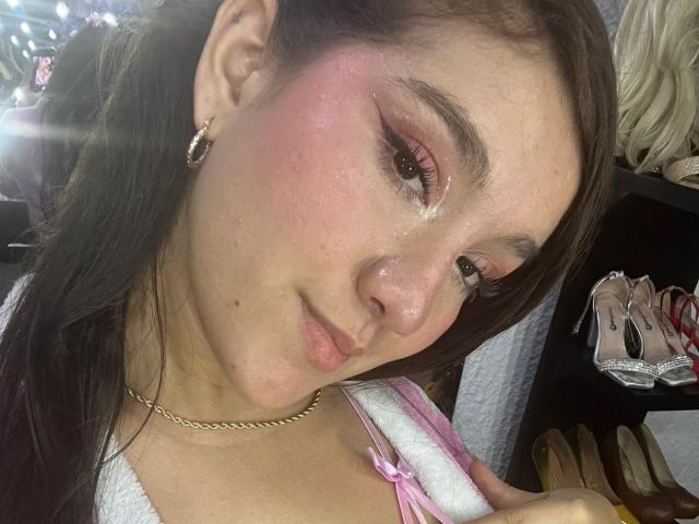 GagingZhara has pink blush on her cheeks, brown eyes with eyeliner, long brown hair, and a gold necklace. Her white top shows a pink strap. Her nose is slightly shiny.