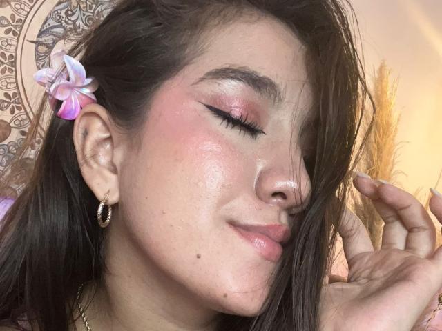 GagingZhara with pink eyeshadow, rosy cheeks, and a small beauty mark on her left cheek, has a pink flower in her brown hair, wearing gold earrings, and touching her hair.