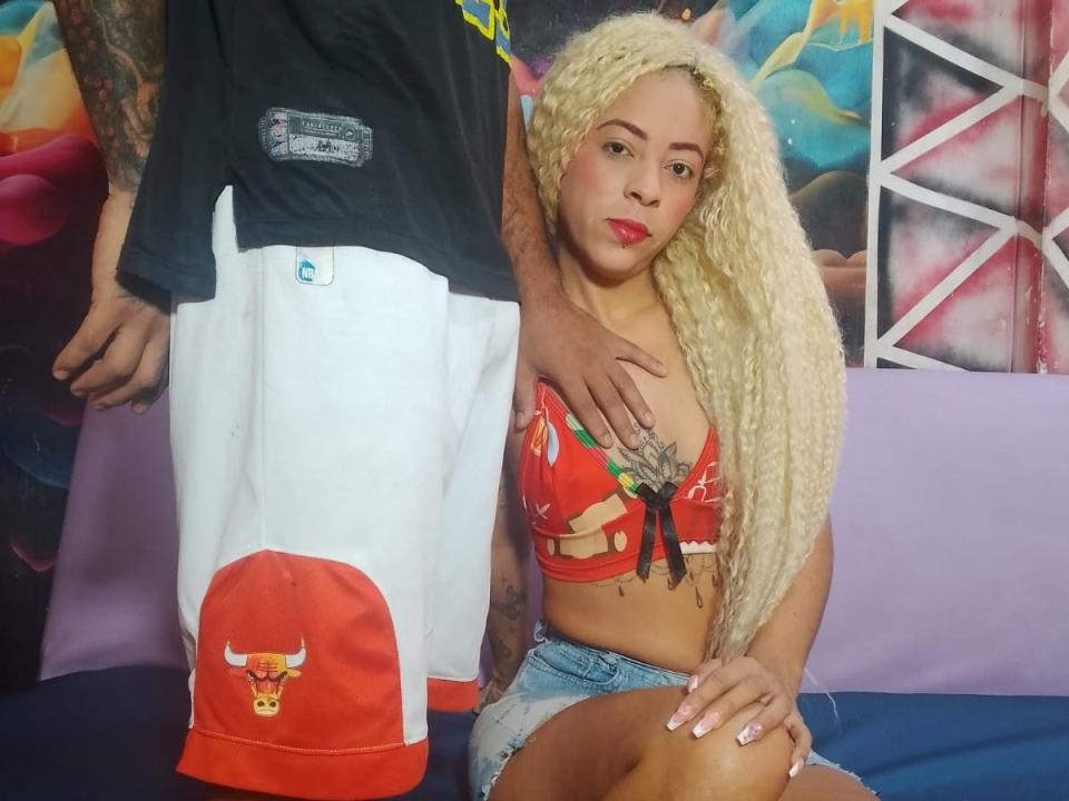 WeAreSeduction with long blonde curls, red crop top, and denim shorts, standing next to a tattooed dude in black shirt and white bull-print shorts, hand on her shoulder.