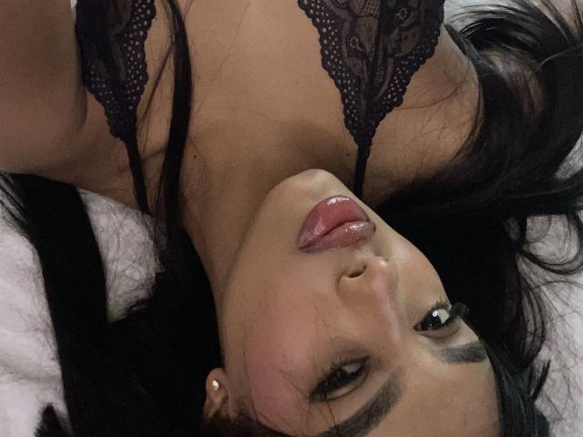 Melli69 with black lace bra, lying down, head tilted, glossy pink lips, dark eyes, and long black hair, wearing small pearl earrings, showing her tits.