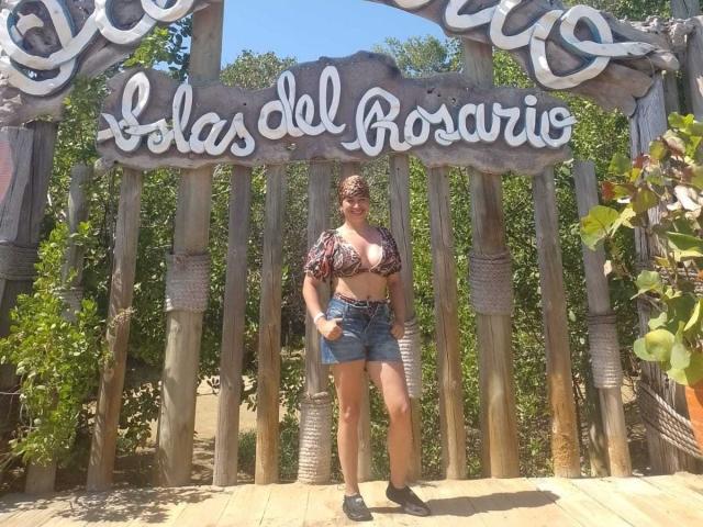 HanaMariana stands in front of a wooden archway with "Las Olas del Rosario" carved in white script. She's a plus-sized woman with dark hair in a bun, wearing sunglasses, a patterned crop top revealing large cleavage, and denim shorts. She has a confident stance, one hand on her hip. The background shows green foliage and a clear blue sky. She's wearing black sneakers. The wooden archway has vertical slats and rope accents. The image has a sunny, outdoor vibe.