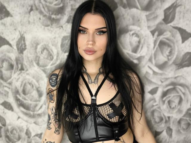 TattooDolly con lunghi capelli neri, che indossa una maglia nera in rete e pelle, che mostra i suoi seni con motivi a stelle nere, braccia tatuate e un trucco completo, in piedi di fronte a un muro con un motivo a rose..