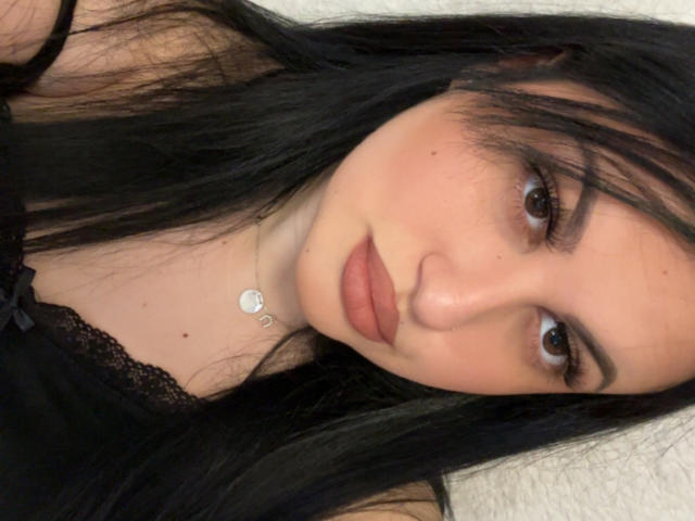 ReddVelvet has long black hair, brown eyes, and full pink lips. She wears a black lace top with a small bow and a silver necklace with a heart charm.