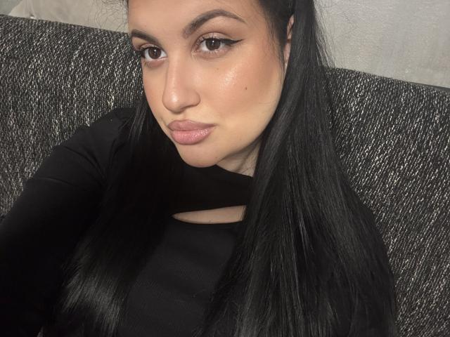 ReddVelvet has long black hair, wearing a black top with a cutout, brown eyes, and pinkish lips, sitting on a gray textured couch.