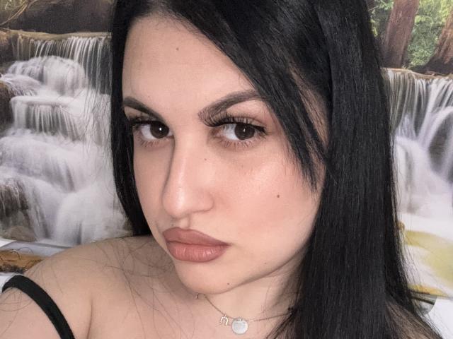 ReddVelvet has black hair, brown eyes, and is wearing nude lipstick. She's looking at the camera with a slight pout, wearing a black strap and a silver necklace.