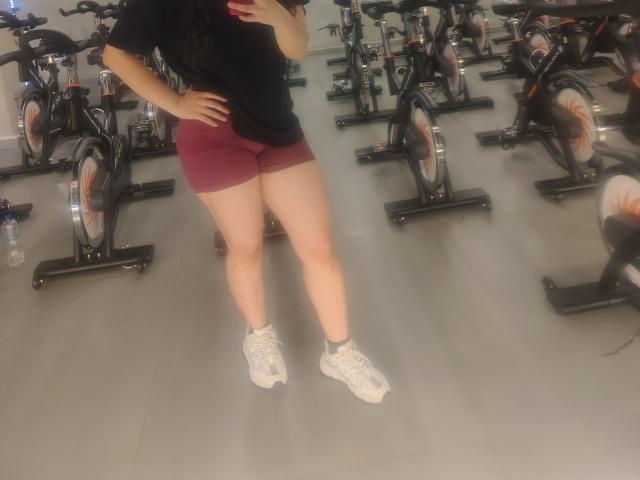 LatinaCulChaud in a gym, wearing a black t-shirt, red shorts, and white sneakers, with her hand on her hip, standing between stationary bikes.