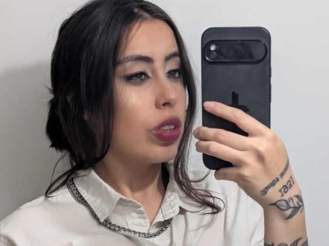 KimyR with dark hair, pink lips, white shirt, holding phone, tattoos on right arm, chain necklace, taking selfie, looking at camera.