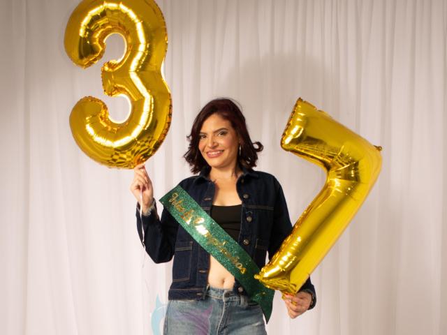 KatteRouse with red hair, smiling, wearing denim jacket, holding gold "3" and "7" balloons, green sash, showing her belly button, white curtain background.