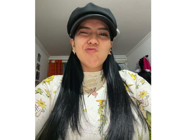 Velka with black pigtails, winking, wearing a black flat cap and white floral shirt, puffy lips, small gold earrings, in a brightly lit room.