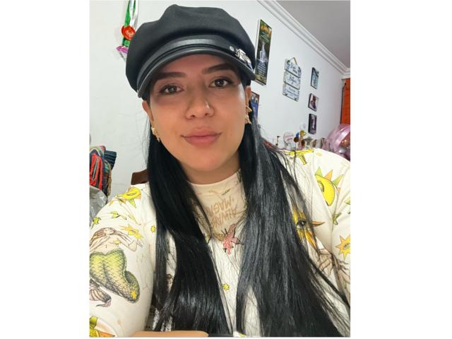 Velka with long black hair, wearing a black cap and yellow star-patterned shirt, has a cute face and a slight smile, showing her nose ring.