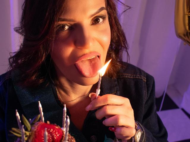 KatteRouse, la langue tirée, léchant une bougie sur un gâteau avec des fraises et des bougies, portant une veste bleu foncé, des cheveux bruns et un collier, dans une lumière chaude..