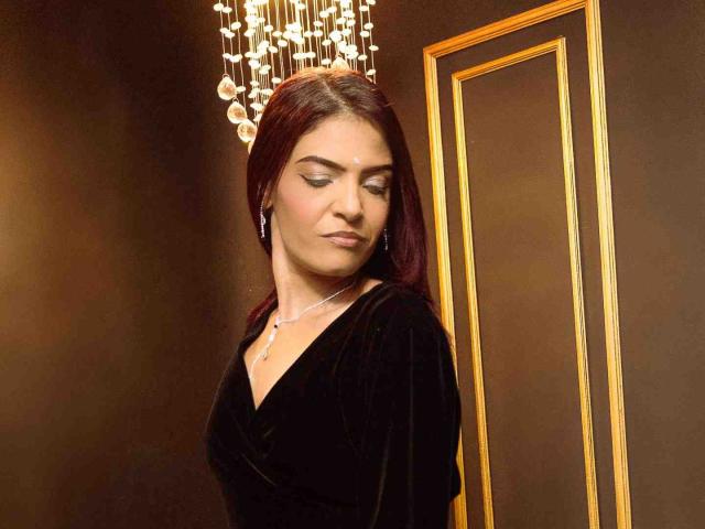 KatteRouse with dark red hair, wearing a black velvet dress, silver necklace, and earrings, standing near a gold-framed mirror, looking down with a slight smile.