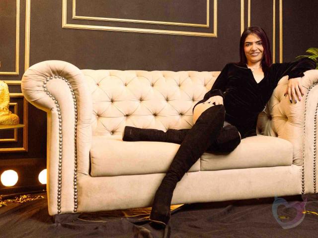 KatteRouse with dark red hair, black velvet top, and black thigh-high boots, sitting on a beige tufted sofa, legs crossed, smiling. Her ass is slightly visible.
