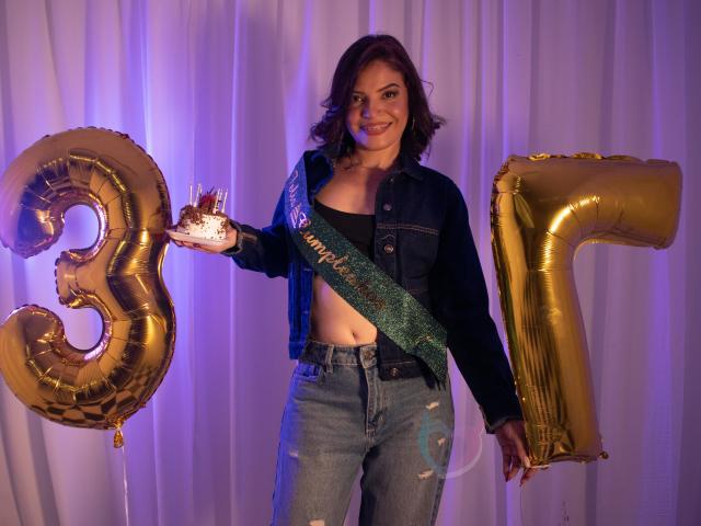 KatteRouse with red hair, denim jacket, black top, and ripped jeans, holding a "3" balloon and golden "7" balloon, sash, and small cake, smiling.
