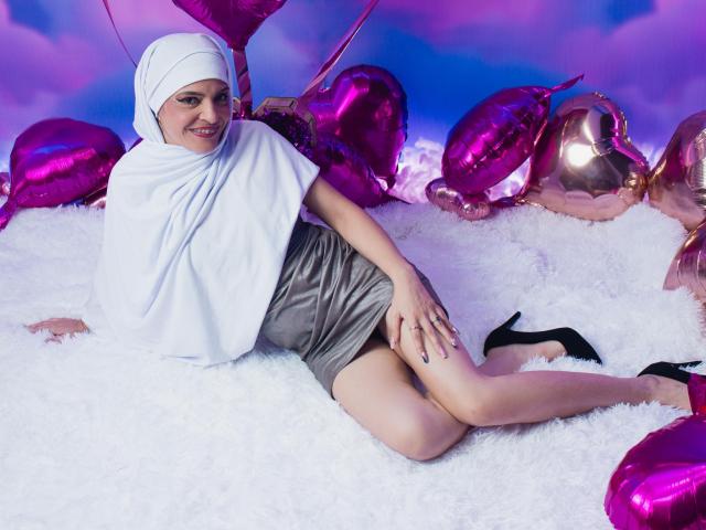 KatteRouse lying on white fluffy carpet, wearing a white hijab, gray mini-dress, and black high heels, with pink heart balloons and a white shawl covering her tits.