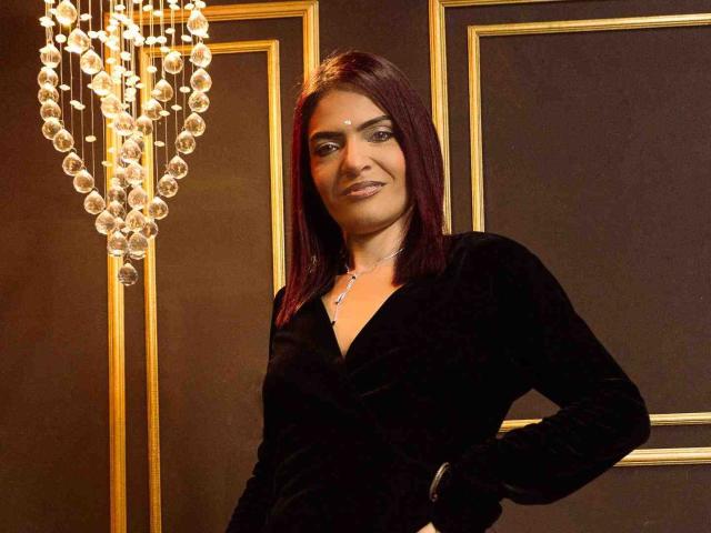 KatteRouse with dark red hair, black velvet dress, and necklace, standing with hand on hip, smiling, chandelier with hanging crystals on left.