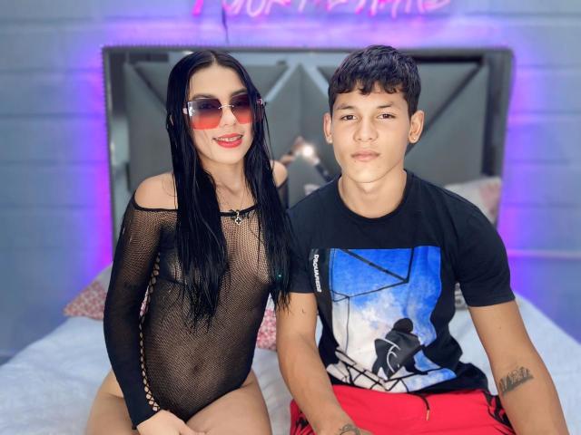 SweetCoupple with long black hair, red sunglasses, and a sheer black bodysuit, showing tits, sits on a bed with a short-haired boy in a black graphic tee.