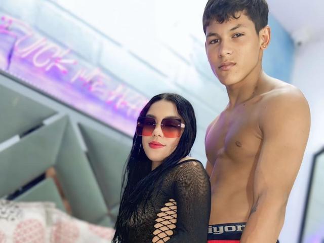 Shirtless SweetCoupple with toned abs and short brown hair stands next to SweetCoupple in a black fishnet dress, red sunglasses, and red lipstick, both looking at the camera.