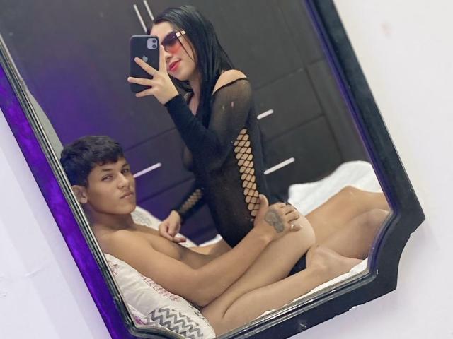 Mirror selfie of SweetCoupple with black fishnet dress, red lipstick, and sunglasses, standing over a naked SweetCoupple with short dark hair and tattoo, lying on a bed.