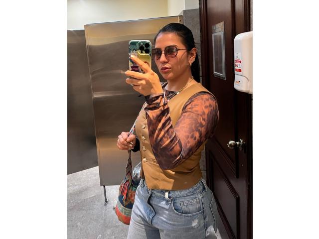 Velka with dark hair, glasses, and brown vest, holding phone in a restroom, wearing blue jeans, puffy brown sleeves, and a colorful bag, taking a mirror selfie.