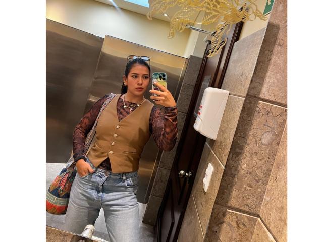 Velka with brown vest, patterned shirt, and blue jeans, holding phone in mirror, has medium tits, black hair in bun, sunglasses on head, standing in bathroom, bag on side.