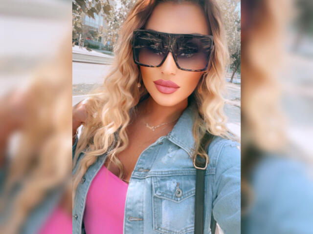 NicolSNaya with blonde wavy hair, big ass, wearing huge black sunglasses, denim jacket, pink top, and pink lipstick, standing in a sunny street.