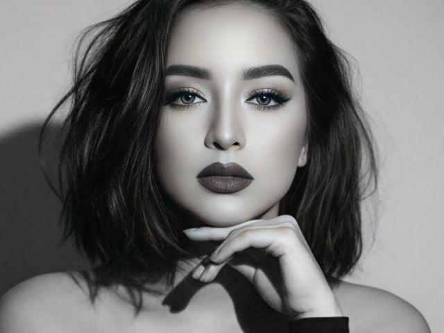 YourDreamGoddess with pale skin, dark wavy hair, green eyes, thick eyebrows, dark lipstick, and a serious expression, touching her chin with her right hand.