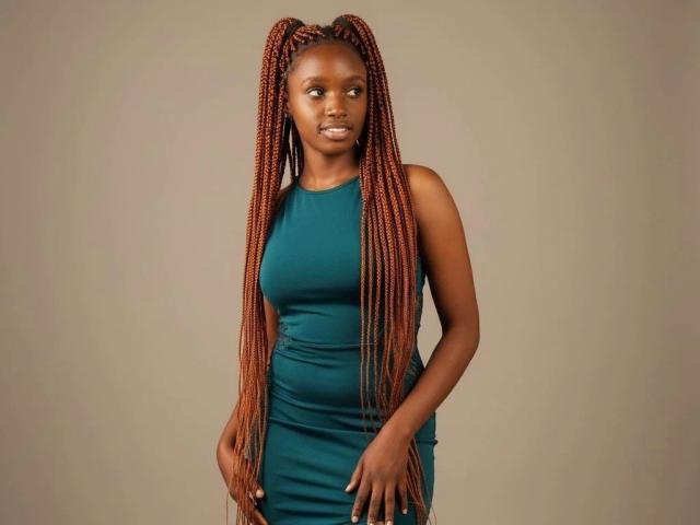 EbonyShy-hot stands centered against a plain beige background. She has long, thick, braided orange-red hair that cascades down past her shoulders. She's wearing a tight, sleeveless teal dress that accentuates her slim, athletic build. Her dark brown skin contrasts with the dress, and she's looking slightly to the right with a subtle, confident smile. Her right hand is relaxed by her side while her left hand rests on her hip. The image is sharp with a focus on her face and hair.