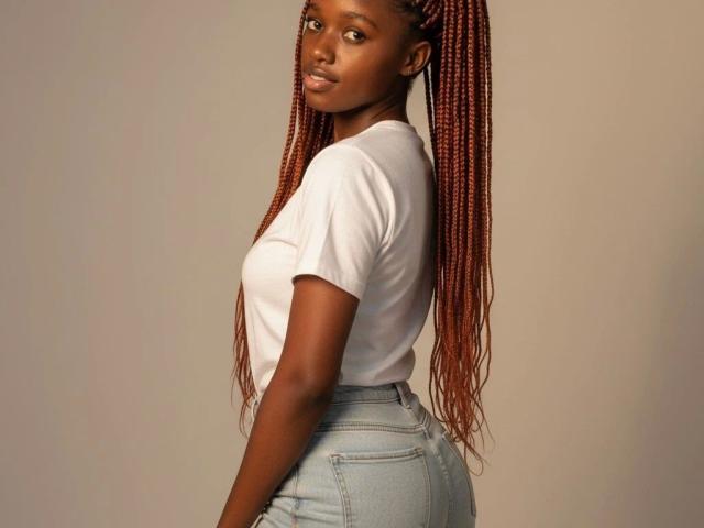 EbonyShy-hot stands facing slightly to the side, looking over her shoulder. She has long, thick, red braided hair extending down her back. She wears a plain white t-shirt and light blue high-waist jeans. Her dark brown skin contrasts with the white shirt. The background is a plain, beige color. Her expression is neutral, lips slightly parted. The image focuses on her upper body, with her right arm partially visible. The composition is simple and centered.