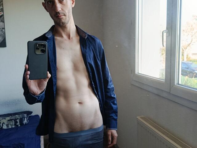 FrancaisH, shirtless, with a lean, muscular torso, blue shirt, gray underwear, holding a phone, mirror selfie, window on the right, daytime, slightly stubbled, serious expression.