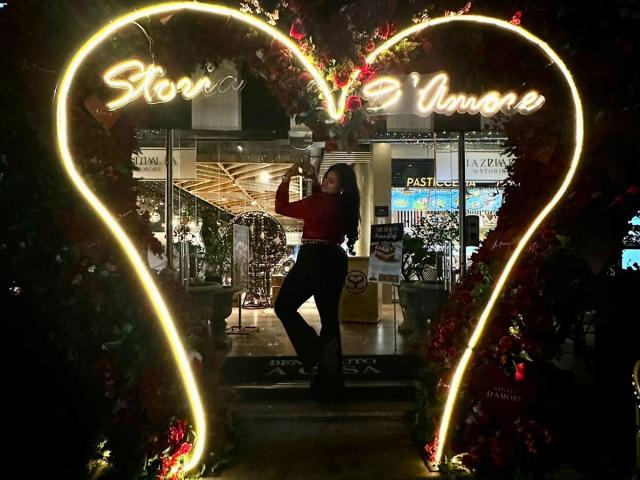 ElyRoss standing in front of a neon heart-shaped sign, wearing a red shirt and black pants, with glowing yellow lights and Christmas decorations, posing with one arm raised.