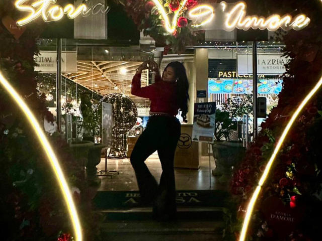 ElyRoss with long dark hair, red top, black pants, standing, holding a wine glass, neon "Storina Vino" sign, Christmas decorations, glowing lights, and a reflective window in the background.