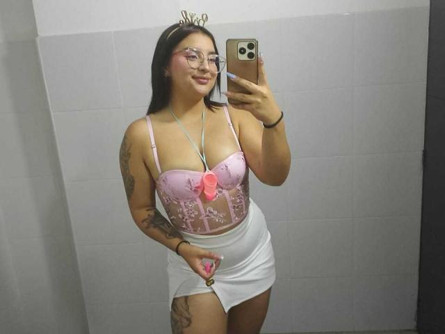 MissSubmissive met een bril, een roze korset met strikjes dat haar borsten laat zien, een witte minirok, een roze ketting, die een telefoon vasthoudt, tatoeages op haar armen, staand in een badkamer, een spiegelzelfportret..