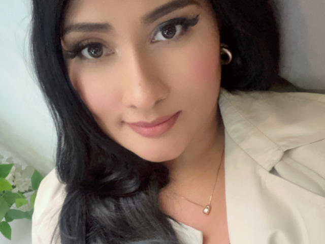 ElyRoss with long black hair, dark eyeliner, and a subtle smile, wearing a white shirt and gold earrings, showing a cute, sexy face with pink blushed cheeks.