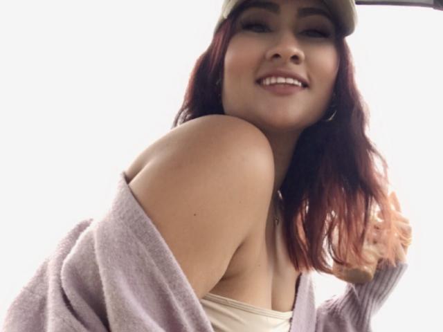 ElyRoss with red hair, smiling, wearing a beige cap, off-shoulder purple sweater, exposing her shoulder and a hint of her tits, against a bright white background.